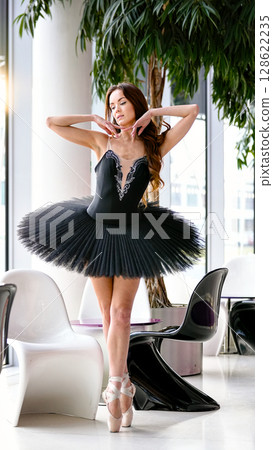Ballet dancer in black tutu posing on pointe near chairs and trees 128622235