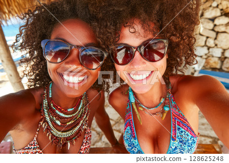 Two smiling women with sunglasses enjoying summer vacation at beach with vibrant atmosphere 128625248