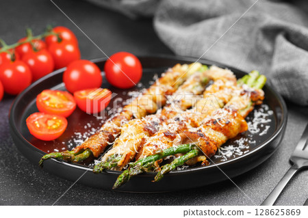 Grilled asparagus wrapped with bacon on black plate with cherry tomatoes on black background Grilled asparagus wrapped with bacon on black plate with cherry tomatoes on black background 128625869