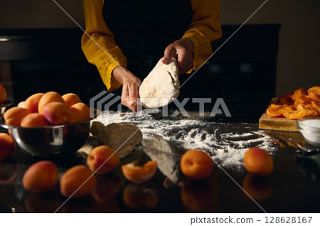 Woman Preparing Dough on a Table Surrounded by Fresh Apricots and Flour in a Rustic Kitchen Setting 128628167
