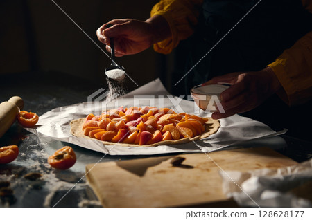 Hands Preparing a Fresh Apricot Tart by Adding Sugar to the Fruit in a Rustic Kitchen Setting Hands Preparing a Fresh Apricot Tart by Adding Sugar to the Fruit in a Rustic Kitchen Setting 128628177