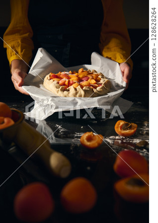 Woman Holding Freshly Baked Apricot Galette on Baking Paper in Warm Kitchen Light 128628264