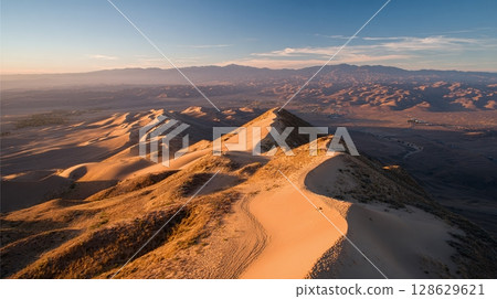 Golden Hour on Desert Dunes: A serene landscape of rippling sand under a clear sky, illuminated by the warm light of sunrise or sunset. Mountains in the distance. 128629621