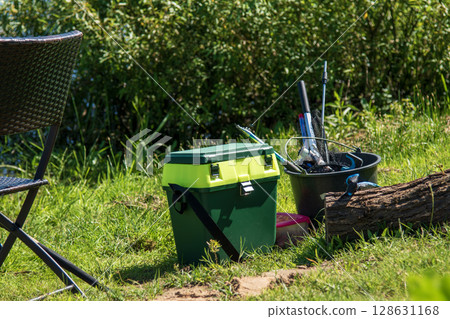 Fishing tackle box and gear on green grass by river with chair and bucket 128631168