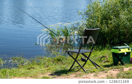 Fishing rod on holder by river with chair and tackle box on green grass Fishing rod on holder by river with chair and tackle box on green grass 128631169