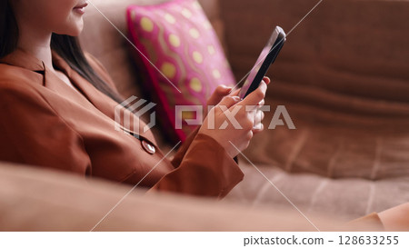 Modern Asian adult woman confidently uses smartphone digital communication comfortable sofa home living Modern Asian adult woman confidently uses smartphone digital communication comfortable sofa home living 128633255