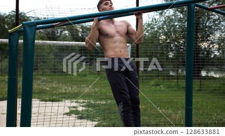 Young muscular sportsman doing pull ups at sports ground. Athletic guy exercising outdoor. Strong sporty man working out at summer nature. Concept of healthy and active lifestyle. Slow motion 128633881
