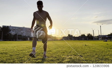 Young man juggling soccer ball on stadium at sunset. Professional footballer kicking ball at green field. Sportsman practicing tricks at meadow with sunlight at background. Freestyle football Young man juggling soccer ball on stadium at sunset. Professional footballer kicking ball at green field. Sportsman practicing tricks at meadow with sunlight at background. Freestyle football 128633906