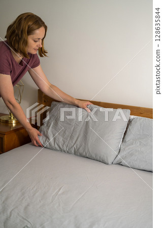 Woman holding a pillow with fresh clean pillowcases, grey cotton bed sheets 128635844