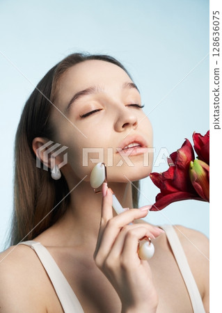 Woman applying skincare with roller while holding a flower in a serene setting 128636075