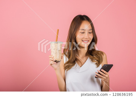 Asian beautiful young woman holding drinking brown sugar flavored tapioca pearl bubble milk tea, Smiling portrait female hold smartphone, studio shot isolated on pink background, milk beverage concept Asian beautiful young woman holding drinking brown sugar flavored tapioca pearl bubble milk tea, Smiling portrait female hold smartphone, studio shot isolated on pink background, milk beverage concept 128636195