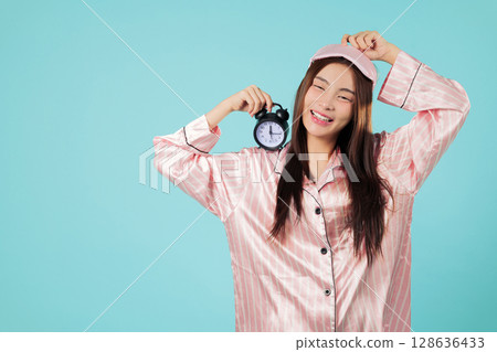 Young asian woman holding alarm clock after waking up from sleep, eye mask on head, wearing pajamas and a yawn betrays she needs more sleep time, headshots isolated studio blue background Young asian woman holding alarm clock after waking up from sleep, eye mask on head, wearing pajamas and a yawn betrays she needs more sleep time, headshots isolated studio blue background 128636433