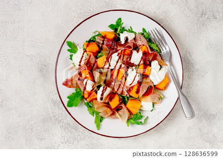 salad with slices of ripe melon, prosciutto, mozzarella and fresh arugula, decorated with balsamic glaze. 128636599