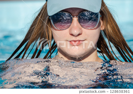 Teenage girl wearing sunglasses and a cap relaxing in a swimming pool, with wet hair and calm expression, enjoying a sunny summer day Teenage girl wearing sunglasses and a cap relaxing in a swimming pool, with wet hair and calm expression, enjoying a sunny summer day 128638361