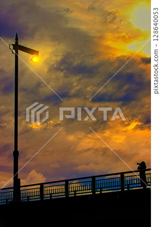 A woman standing on a bridge taking pictures of a street lamp and the sunset 128640053