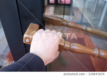 A hand grasping the wooden handle of a door to open a modern entrance to a building A hand grasping the wooden handle of a door to open a modern entrance to a building 128641634