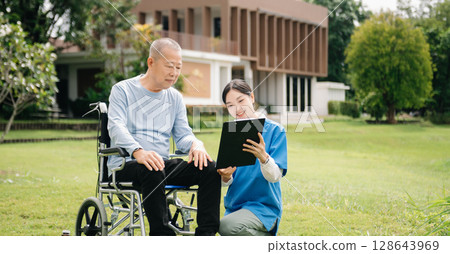 Asian careful caregiver or nurse and the happy patient in a wheelchair are walking in the garden. to help and encourage and rest your mind with green nature. Use a stethoscope to listen Asian careful caregiver or nurse and the happy patient in a wheelchair are walking in the garden. to help and encourage and rest your mind with green nature. Use a stethoscope to listen 128643969