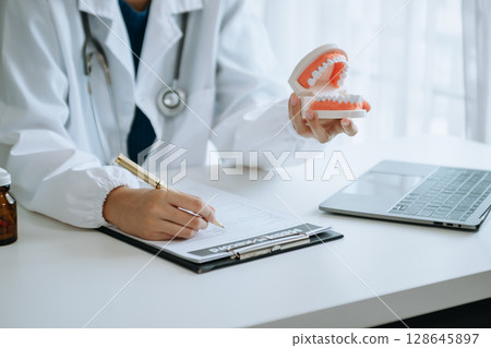 Dentist sitting at table with jaw samples tooth model and working with tablet and laptop in dental office professional dental clinic. 128645897