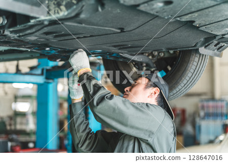 A male auto mechanic performing maintenance, inspection and work under a vehicle going up on a lift 128647016