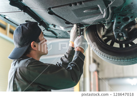 A male auto mechanic performing maintenance, inspection and work under a vehicle going up on a lift 128647018
