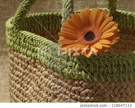 Close up of woven basket with vibrant orange flower inside, showcasing textured straw material and natural earthy tones, evoking warm, rustic, and cozy atmosphere 128647113