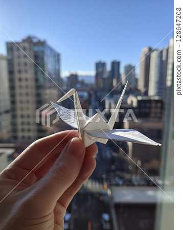 Person holds white origami crane delicately in their hand, filled with hope and aspiration against backdrop of city skyline 128647208