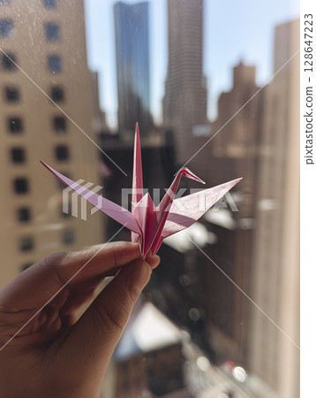 Pink origami crane, delicately held, looks optimistic against cityscape backdrop, evoking sense of peace and hope Pink origami crane, delicately held, looks optimistic against cityscape backdrop, evoking sense of peace and hope 128647223