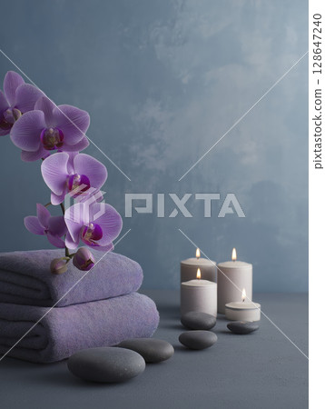 Scene shows stack of purple orchid flowers atop soft towels, lit by candles, creating soothing and peaceful ambiance for relaxation 128647240