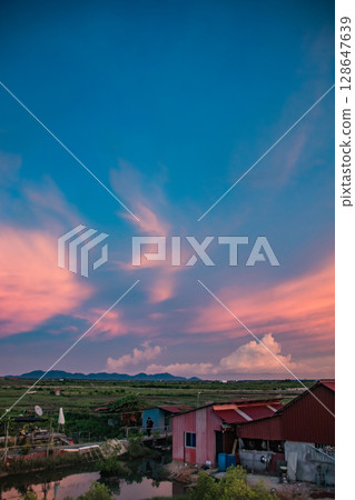 Tranquil summer sunset scene with a colorful sky and distant fields stretching beyond rooftops of tin houses in the rural countryside of Kampot, Cambodia Tranquil summer sunset scene with a colorful sky and distant fields stretching beyond rooftops of tin houses in the rural countryside of Kampot, Cambodia 128647639