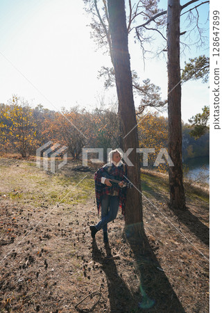 Beautiful blonde girl playing guitar ukulele and singing relaxing enjoying outdoors on sunny autumn day. Hippie woman in boho style training playing musical instrument. Fall leaves 128647899