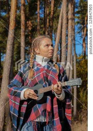 Beautiful blonde girl playing guitar ukulele and singing relaxing enjoying outdoors on sunny autumn day. Hippie woman in boho style training playing musical instrument. Fall leaves 128647903
