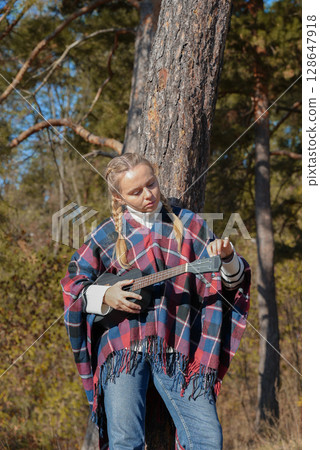 Blonde woman playing guitar ukulele outdoors in autumn forest. Concept of sound therapy, mental health and wellness rituals. Calmness tranquility audio-sensory practices. Aura farming Blonde woman playing guitar ukulele outdoors in autumn forest. Concept of sound therapy, mental health and wellness rituals. Calmness tranquility audio-sensory practices. Aura farming 128647918