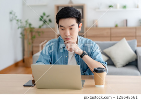 Happy young Asian man working on laptop in living room Happy young Asian man working on laptop in living room 128648561