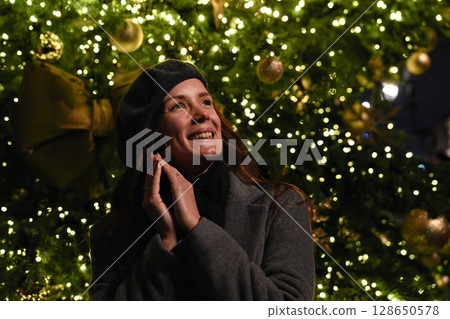 Smiling woman enjoying glowing lights of decorated Christmas tree 128650578