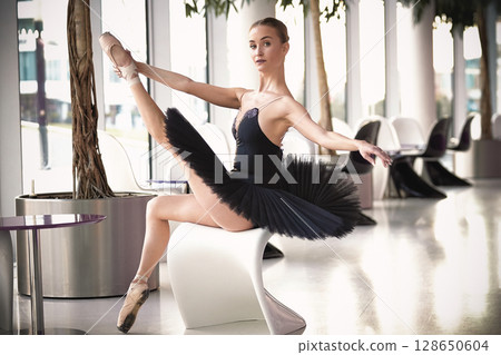 Ballerina in black tutu performs high leg extension on chair indoors Ballerina in black tutu performs high leg extension on chair indoors 128650604