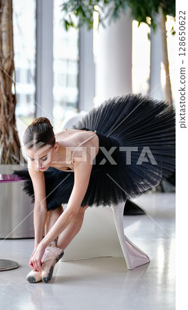 Ballerina in black tutu tying pointe shoes while sitting on modern chair 128650622