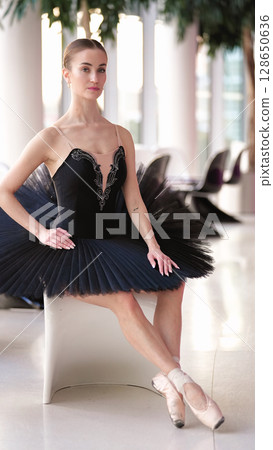 Elegant ballerina in black tutu sitting gracefully on white chair indoors Elegant ballerina in black tutu sitting gracefully on white chair indoors 128650636