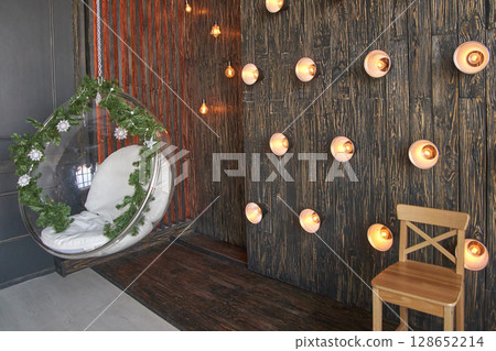 Cozy indoor lounge with hanging chair and string lights on rustic wooden wall 128652214