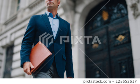 Confident Businessman Walking Outdoors with Briefcase in Urban Environment Confident Businessman Walking Outdoors with Briefcase in Urban Environment 128652617