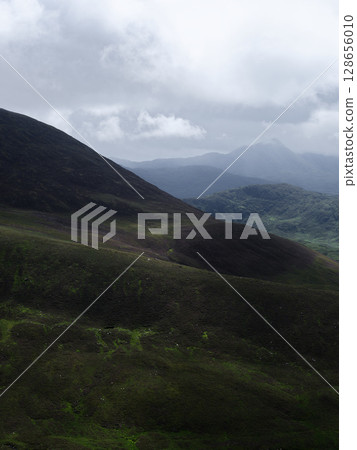 Mountains covered in green vegetation recede into the distance. The sky is grey with cloud cover over the scenic landscape of the Coomloughra Horseshoe in County Kerry. 128656010