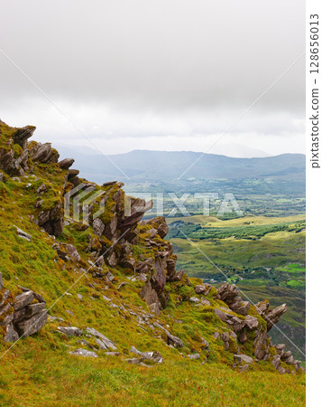 A rocky, grassy slope leads to a vast valley and mountains beyond. The weather is overcast in this scenic view from the Coomloughra Horseshoe trail in Ireland. A rocky, grassy slope leads to a vast valley and mountains beyond. The weather is overcast in this scenic view from the Coomloughra Horseshoe trail in Ireland. 128656013