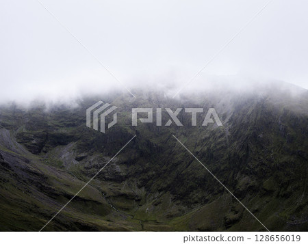 A mountain face is partially obscured by low-hanging fog or mist. Green slopes and craggy rock formations create a dramatic texture under a white sky. 128656019