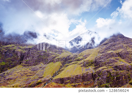Scenic view of Carrauntoohil mountain with green hills, a small waterfall, and fog in County Kerry, Ireland, during the daytime. Scenic view of Carrauntoohil mountain with green hills, a small waterfall, and fog in County Kerry, Ireland, during the daytime. 128656024