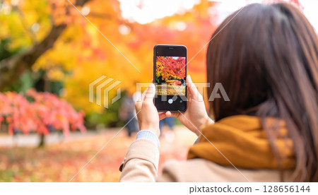 AI Image: A young woman taking pictures of autumn leaves with her smartphone 128656144
