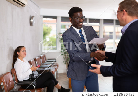 Middle-aged man handshaking with other person in waiting room Middle-aged man handshaking with other person in waiting room 128656455