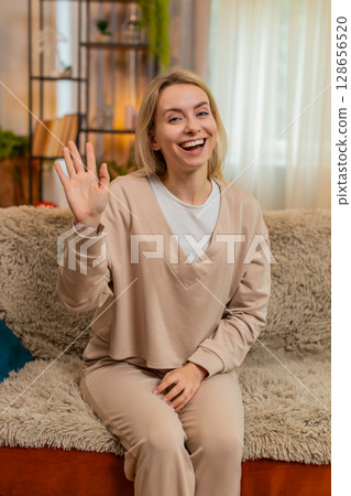 Adult woman smiling and waving hand at home, greeting someone with joy and cheerful energy on sofa 128656520