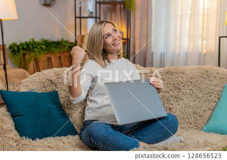 Adult woman celebrating at home after completing work on laptop, raising arm with joy satisfaction 128656523