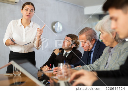 Clever young female making report while office workers of different ages using notebooks at table in office 128656524