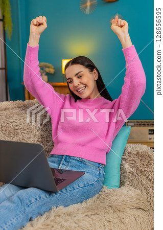 Young woman on sofa raising hands up joyfully celebrating successful finished project on laptop 128656595