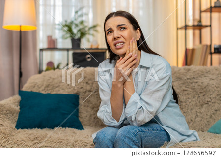 Young woman sitting on sofa holding cheek suffering from strong toothache feeling pain uncomfortable 128656597